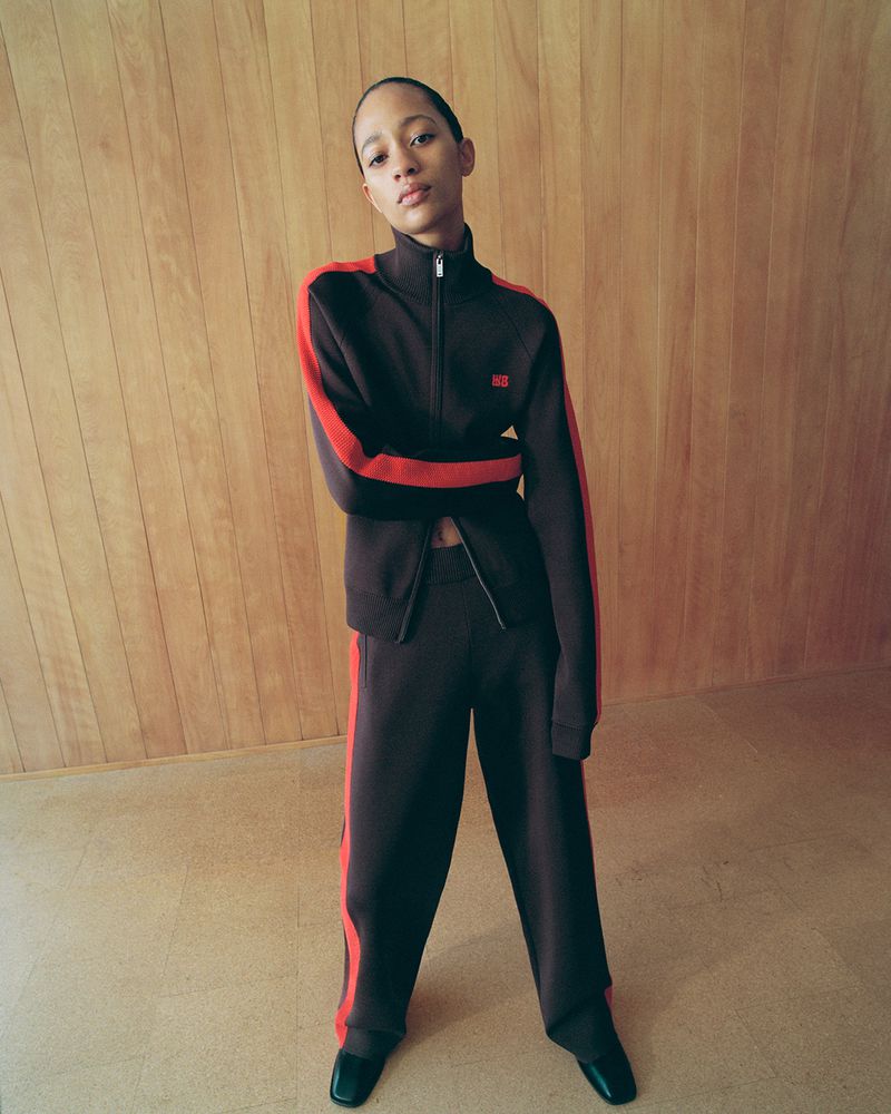 Woman in a brown tracksuit with red stripes posing in front of a wood paneled wall and light beige floor in a studio setting.