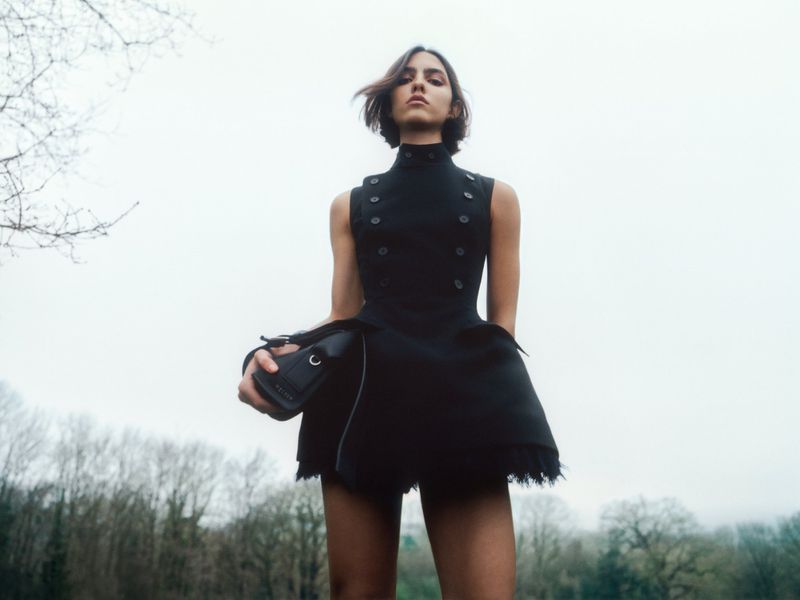 Woman in a short, military-style black dress stands in a park, holding a small black purse. Low-angle shot with trees in the background.