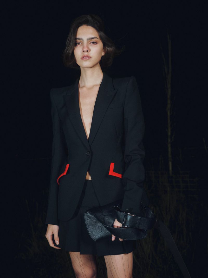 Model in a black blazer with red pocket accents wearing a black skirt against a dark outdoor night background