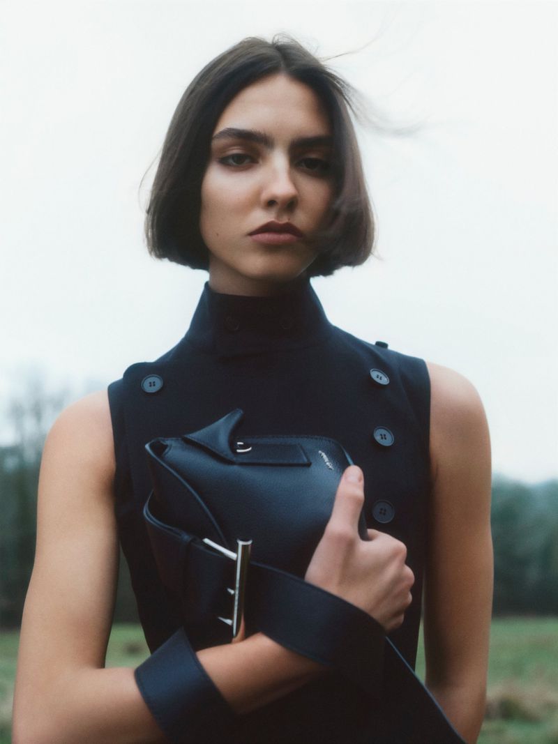 Portrait of a woman in a black sleeveless turtleneck holding a black purse with metal details, posing outdoors in a field.