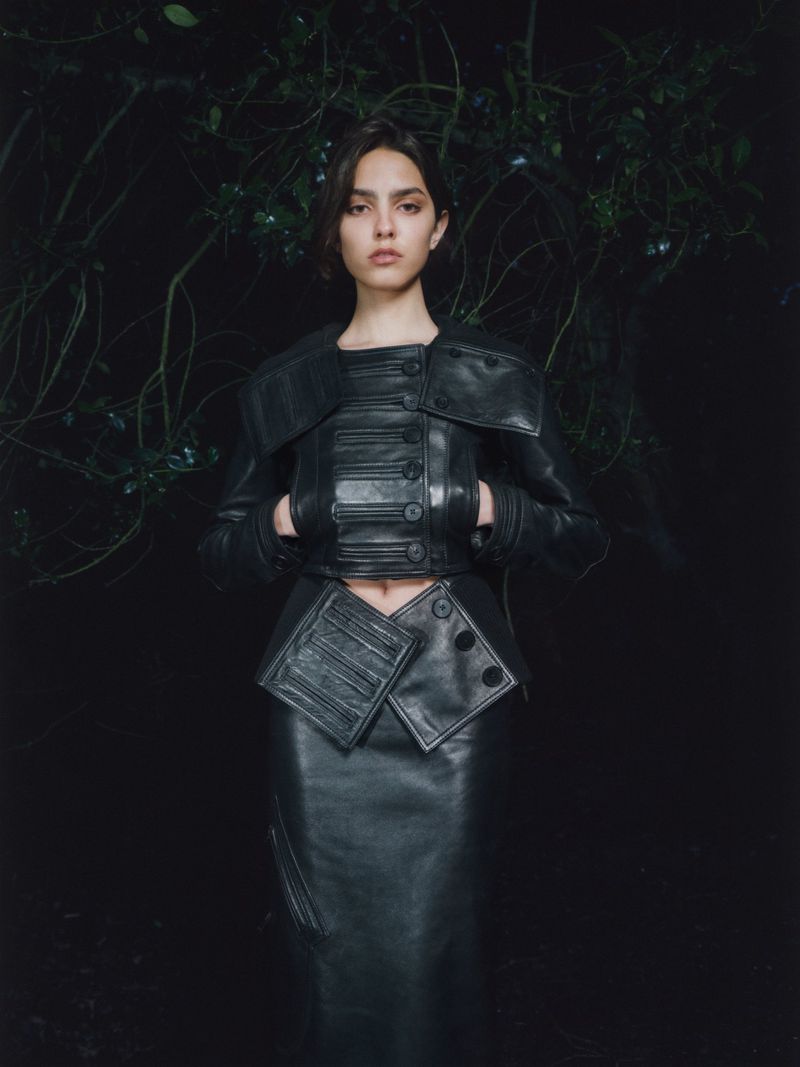 Portrait of a woman in a black leather jacket and skirt posing in front of dark foliage in dim lighting. Fashion editorial.