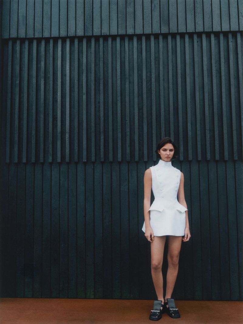 Full-length shot of a woman in a white dress posing against a dark blue slat wall, creating a high fashion editorial image.