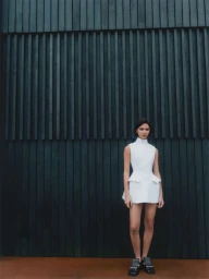 Model wearing a white structured mini dress standing against a dark wooden slatted wall for Net-A-Porter