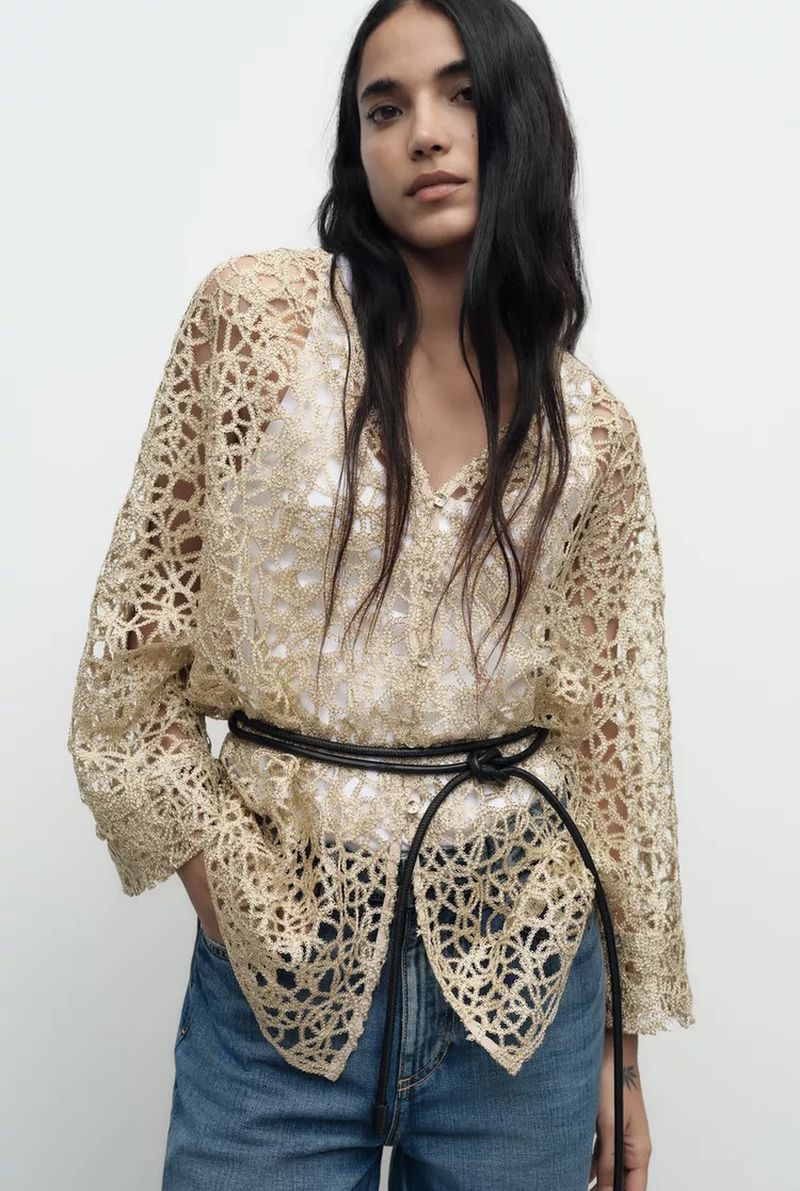Model wearing a cream open-work lace cardigan and blue jeans belted with a thin black cord against a white backdrop.