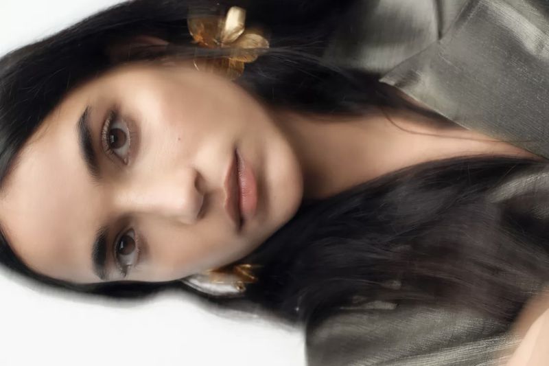 Close-up portrait of an adult woman with dark hair and golden flower earrings, wearing a gray shiny top against a white background.