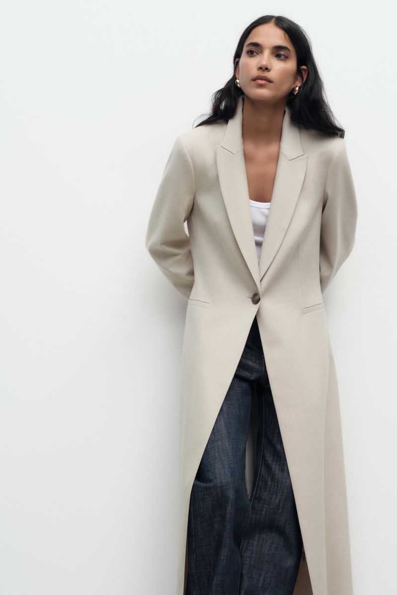 A woman wears a long beige coat, a white shirt and jeans, posing against a white wall in a full shot, minimalist fashion.