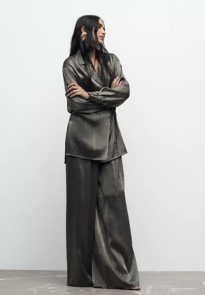 Elegant woman in a satin wrap top and wide-leg pants poses in a studio, showcasing high-fashion style with a sophisticated, modern edge.