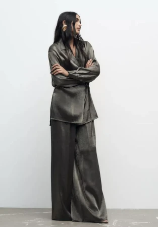 Model with long dark hair wearing a silky olive green matching top and wide-leg trousers standing against a seamless white background.