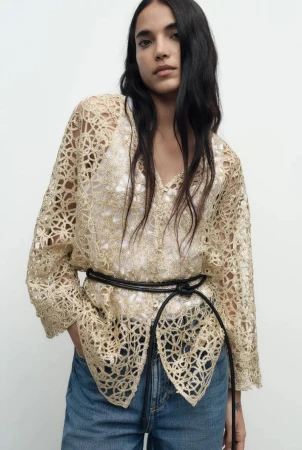 Model wearing a cream open-work lace cardigan and blue jeans belted with a thin black cord against a white backdrop.