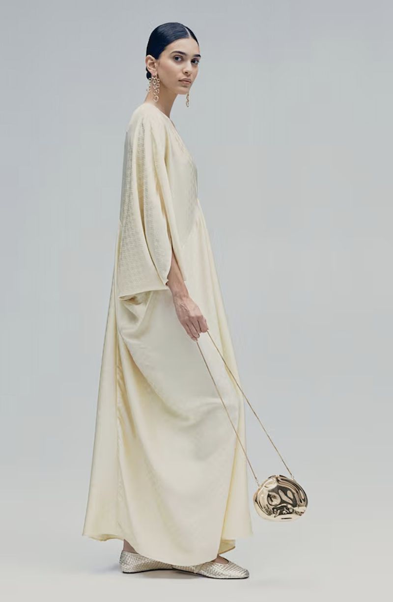 A woman in a flowing cream-colored gown holds a metallic gold sphere-shaped purse against a plain neutral background in a fashion shot.