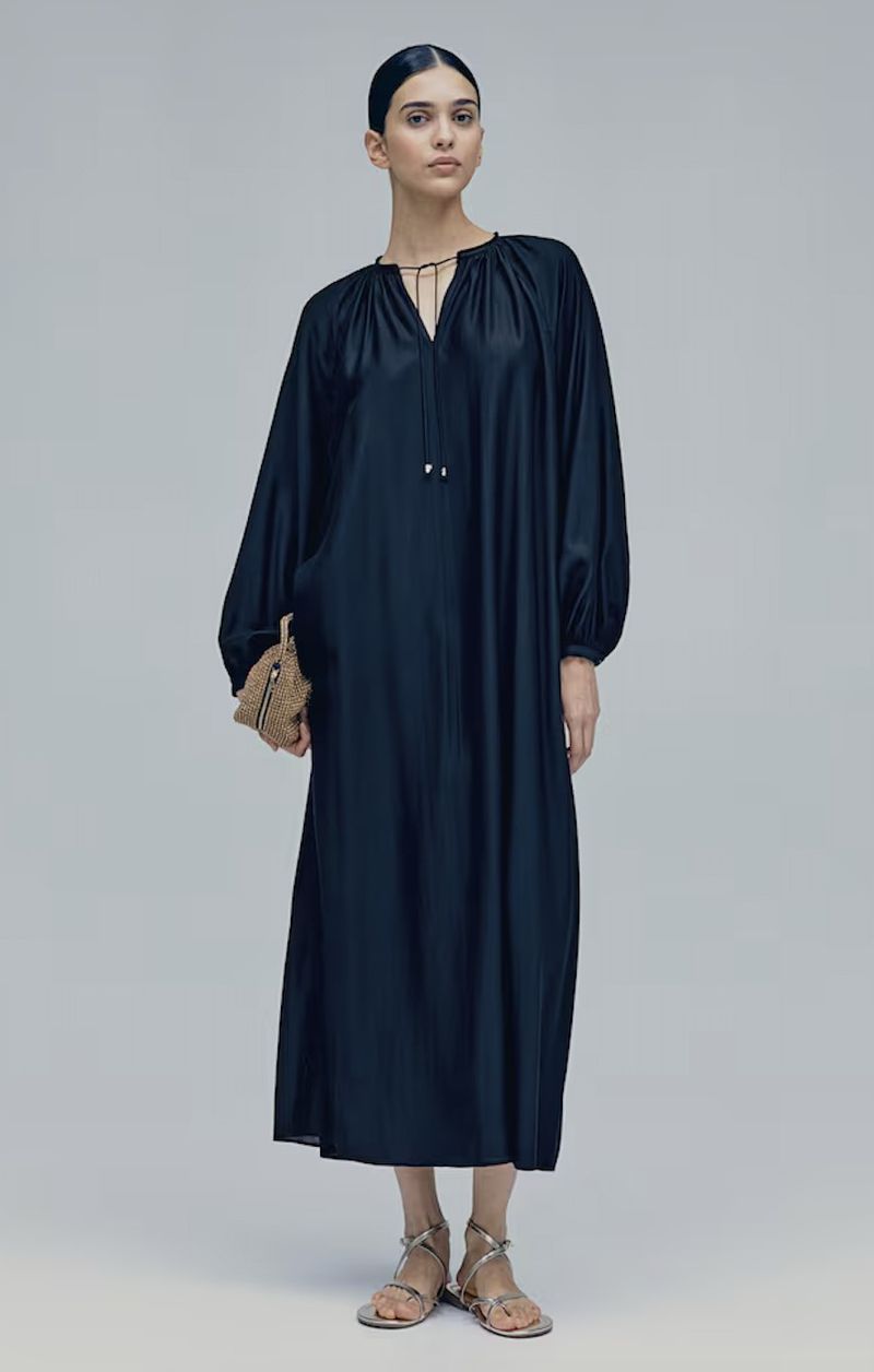 Model wearing a dark long-sleeved midi dress holding a small textured handbag against a plain grey seamless background.
