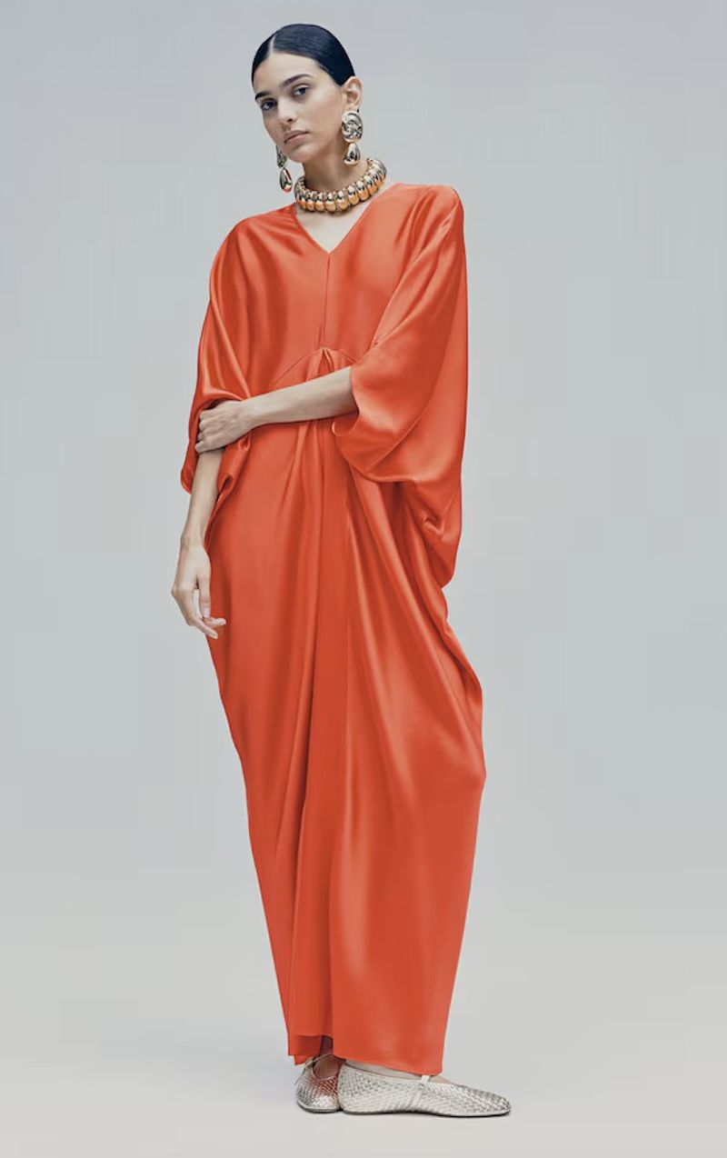 A model poses in a vibrant orange, full-length kaftan dress with statement jewelry, set against a neutral background in a fashion editorial.
