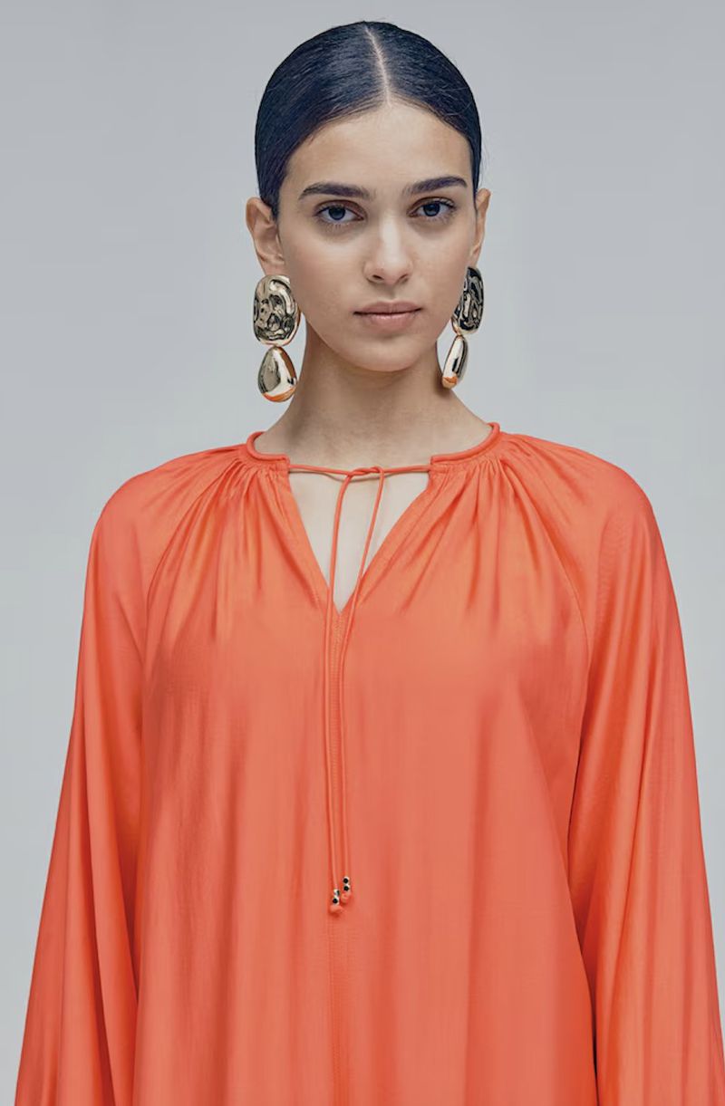 Close-up of a dark-haired woman in an orange, long-sleeved dress with large gold earrings against a light gray studio background.