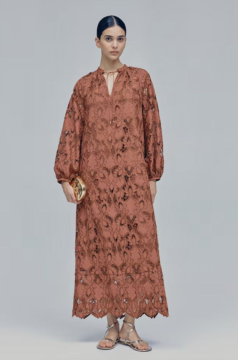 Woman in a flowing terra-cotta maxi dress with a delicate floral pattern and long balloon sleeves, holding a gold clutch.