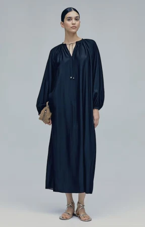 Model wearing a dark long-sleeved midi dress holding a small textured handbag against a plain grey seamless background.