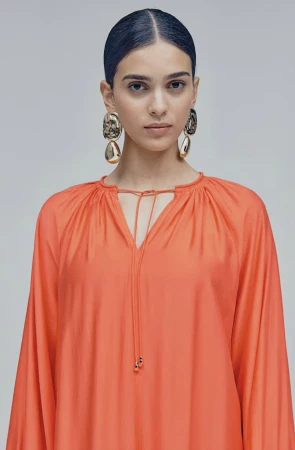 Model wearing an orange gathered blouse and large gold earrings against a plain grey backdrop.