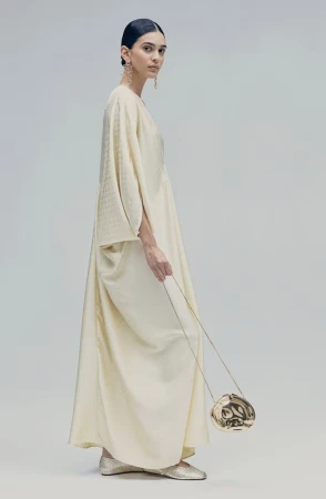 Model in flowing cream kaftan dress holding a metallic gold pouch against a neutral grey studio background