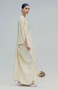 Model in flowing cream kaftan dress holding a metallic gold pouch against a neutral grey studio background