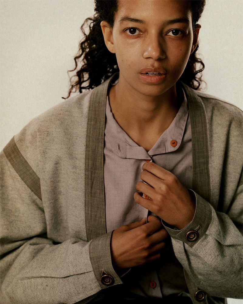 Portrait of a young adult female with curly dark hair, wearing a light purple button-down shirt and a grey, lined cardigan in a studio setting.