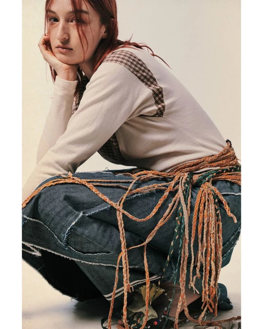 Young woman with auburn hair poses in a photo studio wearing a long-sleeved shirt, denim skirt, and braided rope belt.