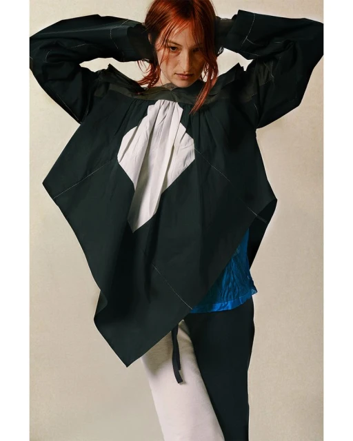A red-haired model showcases a black and white oversized fashion top with an angular design paired with matching pants in a studio setting.