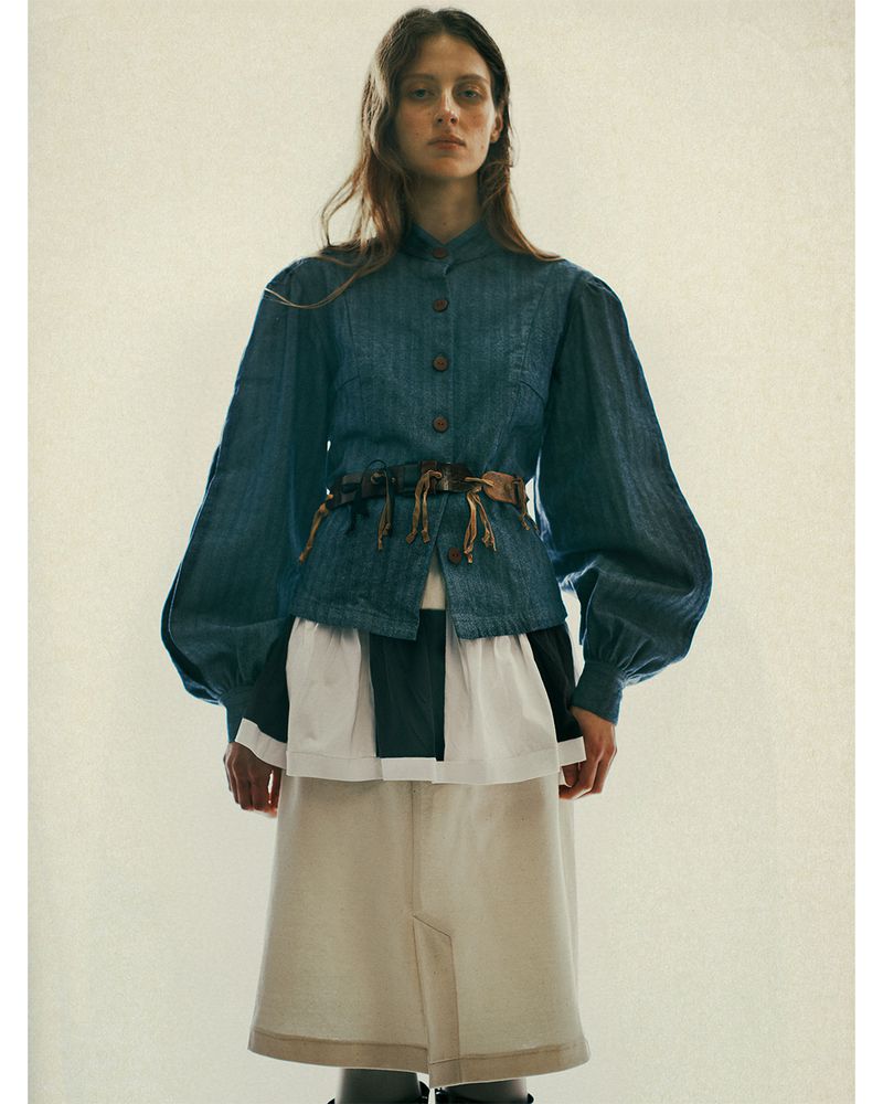 A female fashion model poses in a denim jacket cinched at the waist with a brown belt, a tiered skirt, and dark tights against an off-white backdrop.