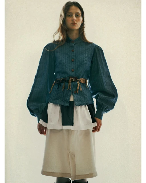 A female fashion model poses in a denim jacket cinched at the waist with a brown belt, a tiered skirt, and dark tights against an off-white backdrop.