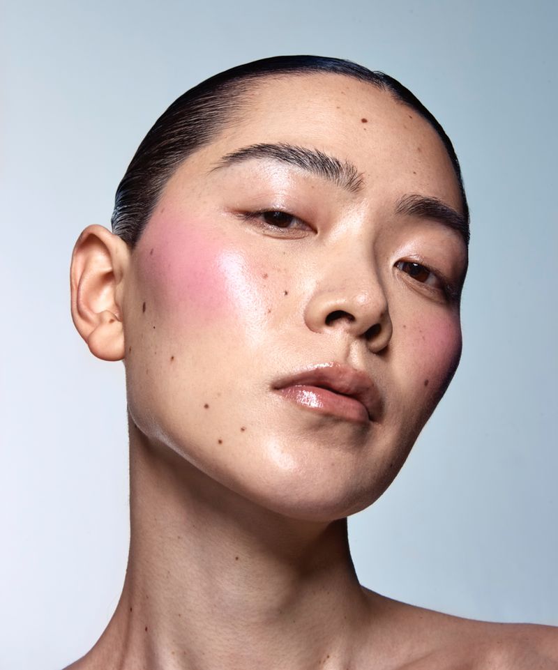 Close-up studio shot of an Asian female adult with glossy skin, pink blush, and wet-look hair against a light blue background.