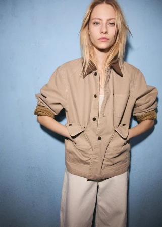 Model wearing a tan utility jacket with a brown collar and white pants against a textured blue wall.