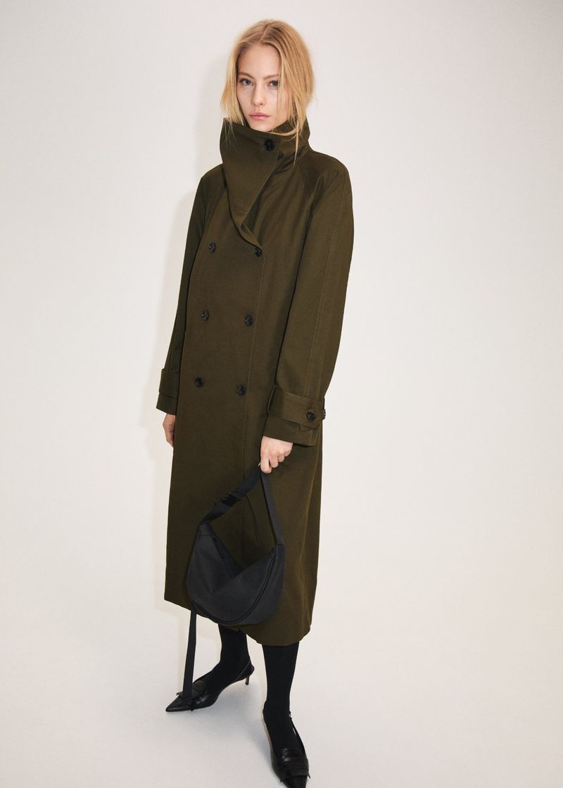 Full-length shot of a woman wearing a double-breasted olive coat, black hobo bag, black stockings, and pointed shoes against a white backdrop.