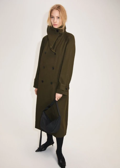 Full-length shot of a woman wearing a double-breasted olive coat, black hobo bag, black stockings, and pointed shoes against a white backdrop.
