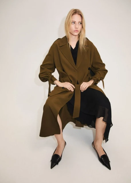 A blonde woman wearing an olive trench coat and black dress is sitting against a white background in a fashion shot, medium angle.