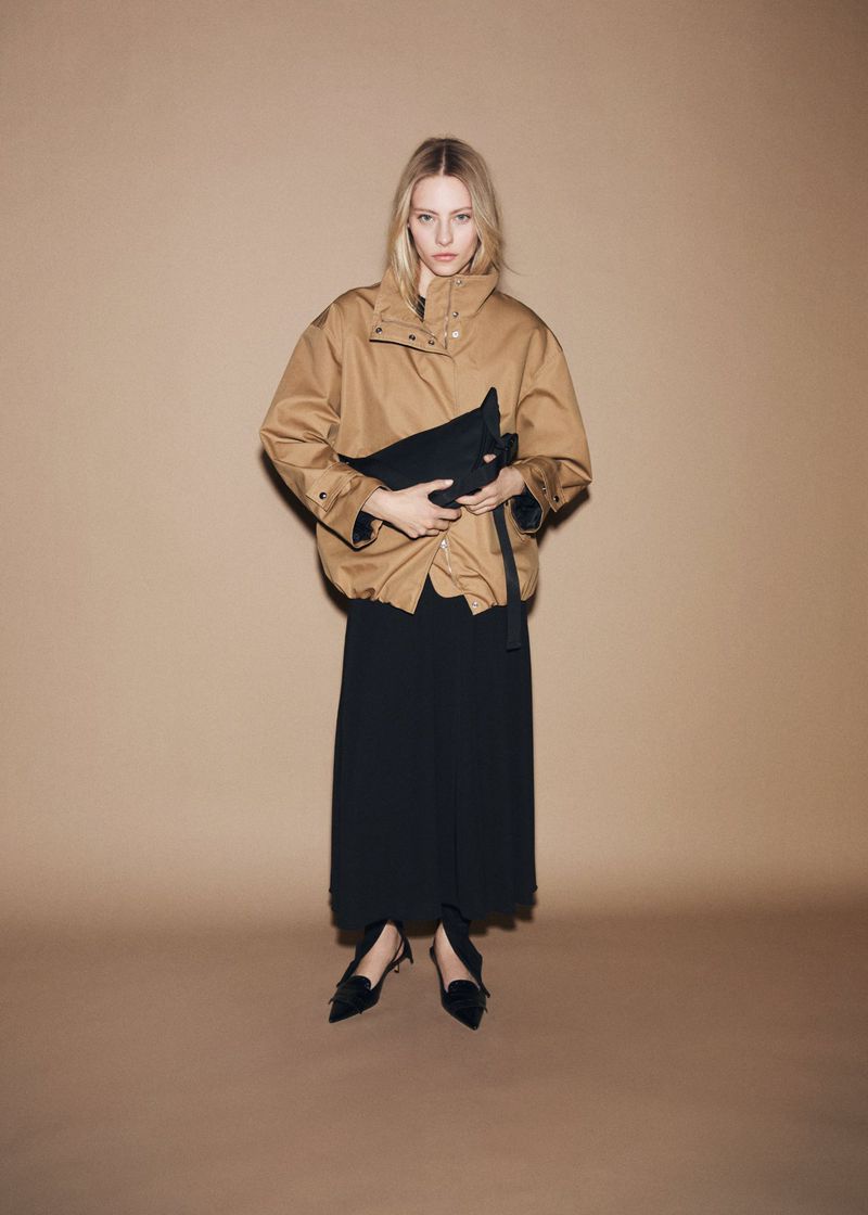 Model wearing a tan jacket and black skirt holding a dark pouch against a neutral brown studio backdrop.