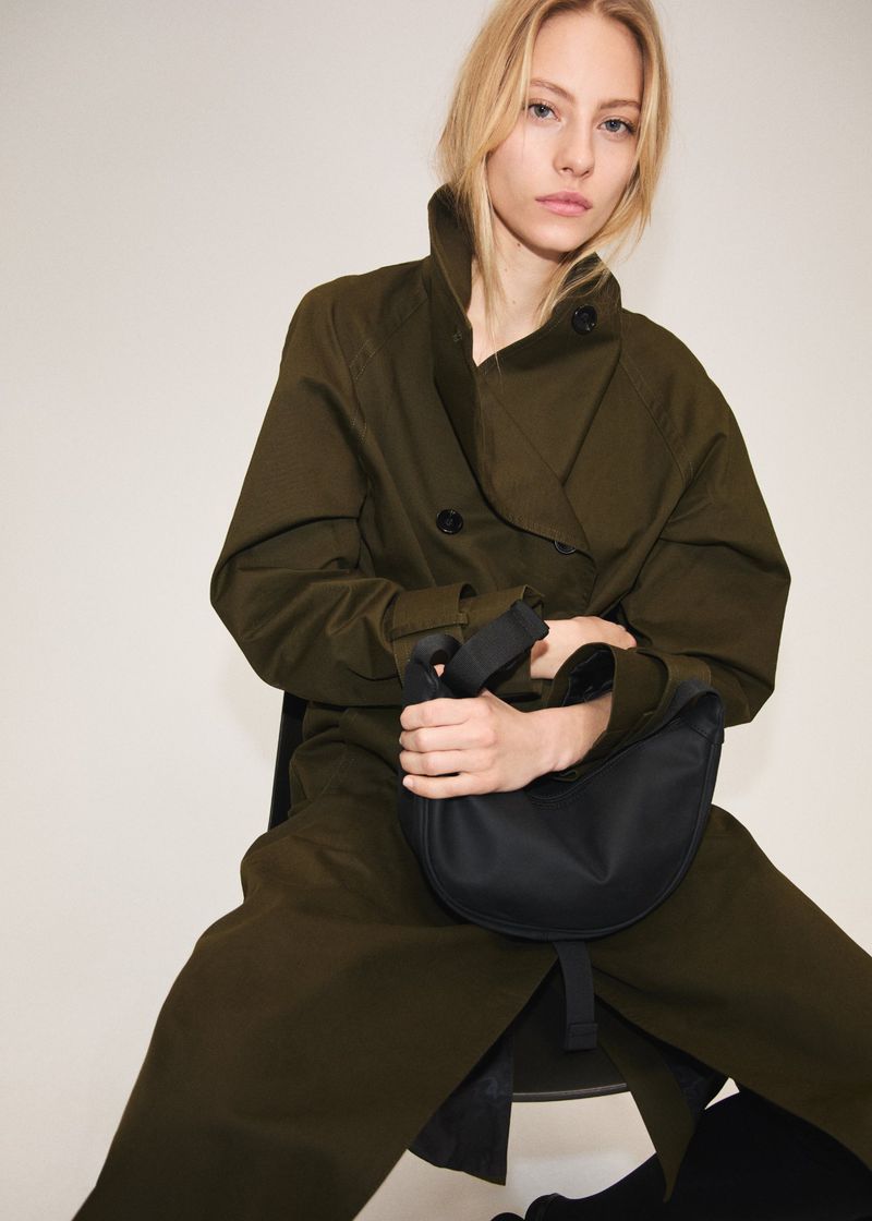 Model Annie Arnander wearing an olive green coat holding a black handbag against a white studio background.