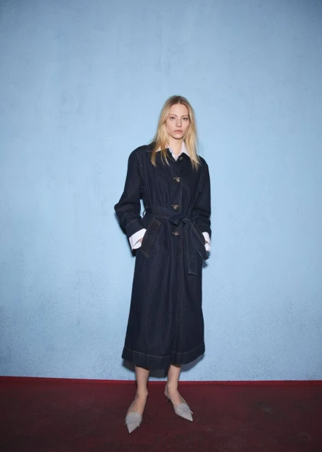 Full-length portrait of a fair-skinned blonde woman in a dark denim trench coat and light-colored flats, posing against a blue wall.