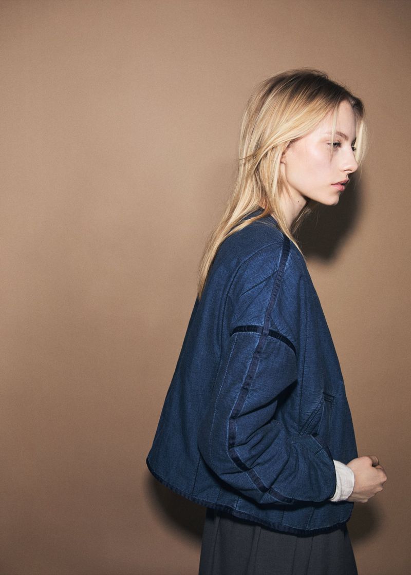 Profile shot of a woman with long blonde hair wearing a blue denim jacket in a studio with a warm beige background, minimalist fashion.
