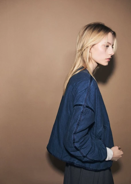 Profile shot of a woman with long blonde hair wearing a blue denim jacket in a studio with a warm beige background, minimalist fashion.