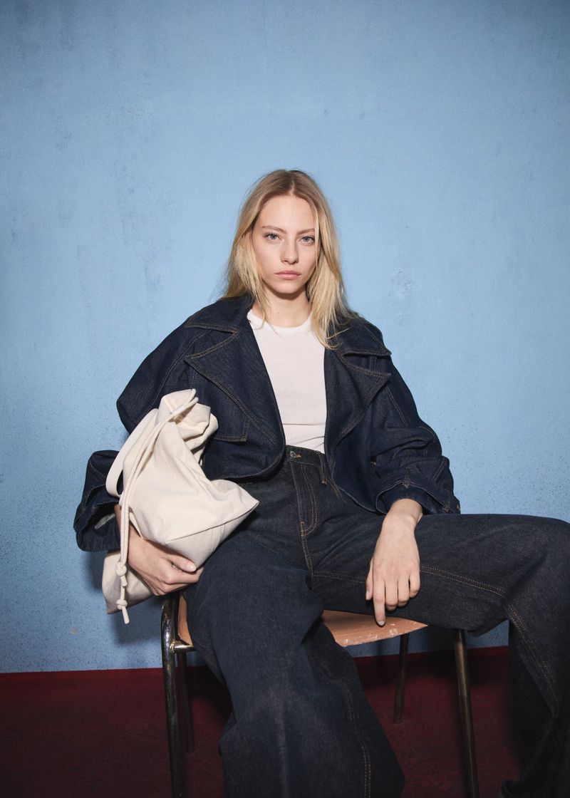 A blond woman in a denim jacket and wide-leg jeans sits on a metal chair, holding a cream-colored bag against a blue backdrop.