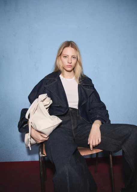 A blond woman in a denim jacket and wide-leg jeans sits on a metal chair, holding a cream-colored bag against a blue backdrop.