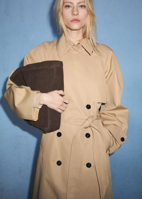 Blonde woman wearing a tan trench coat holds a brown clutch against a blue background in a studio fashion shot. Focus on the woman.