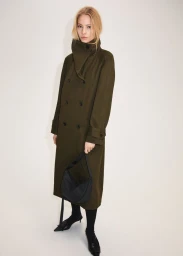 Model Annie Arnander wearing an olive green coat and holding a black handbag against a light studio backdrop.