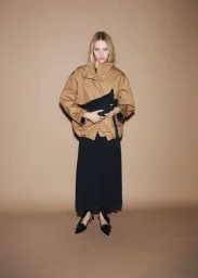 Model wearing a tan jacket and black skirt holding a dark pouch against a neutral brown studio backdrop.