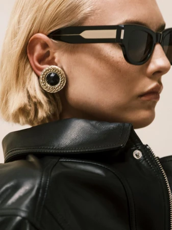 Close-up profile of woman wearing black sunglasses, large gold earring, and black leather jacket against a light background.