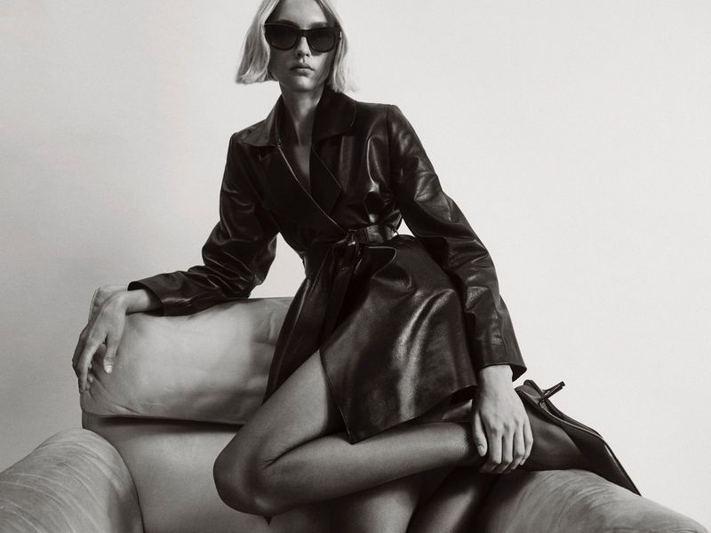 Model in black leather trench coat and sunglasses seated on an armchair against a plain studio background.