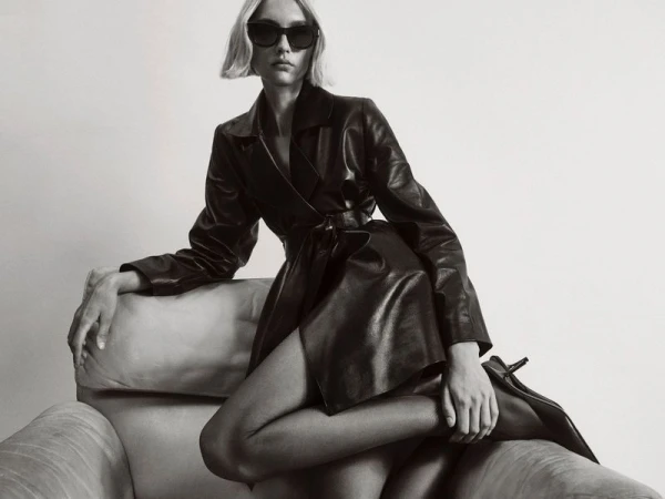 Model in black leather trench coat and sunglasses seated on an armchair against a plain studio background.