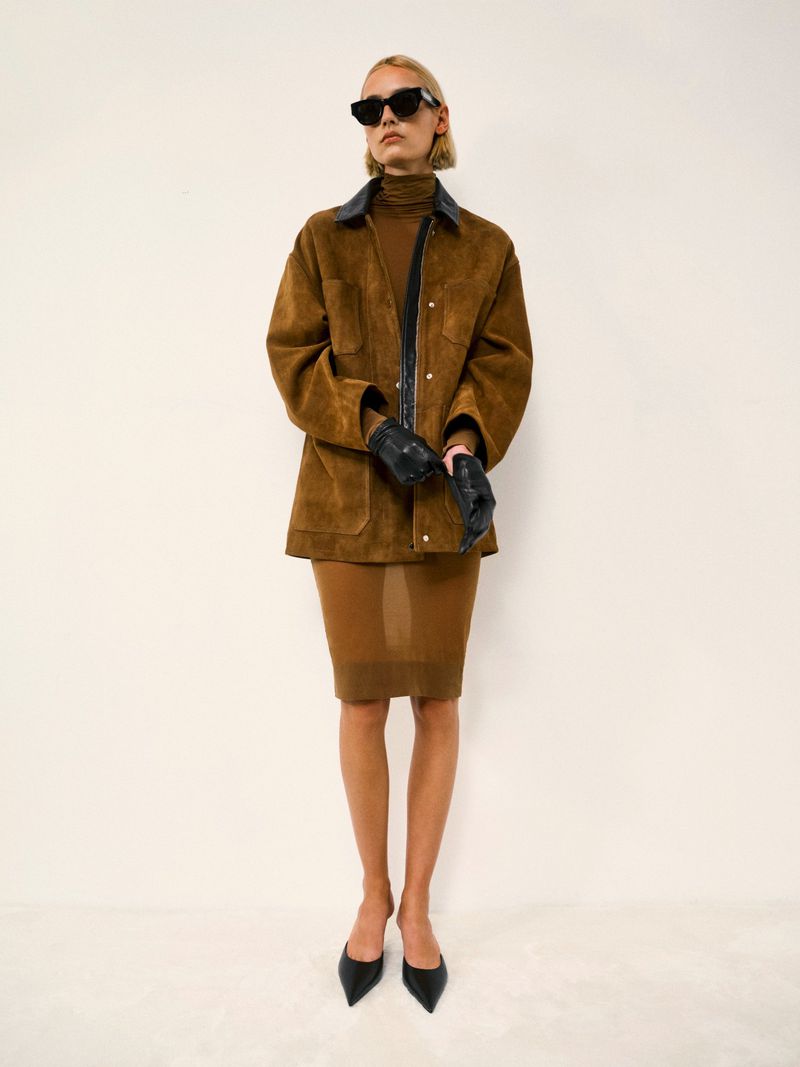 A single adult female poses in a brown jacket, brown skirt, black gloves, and sunglasses against a white background in a studio shot.