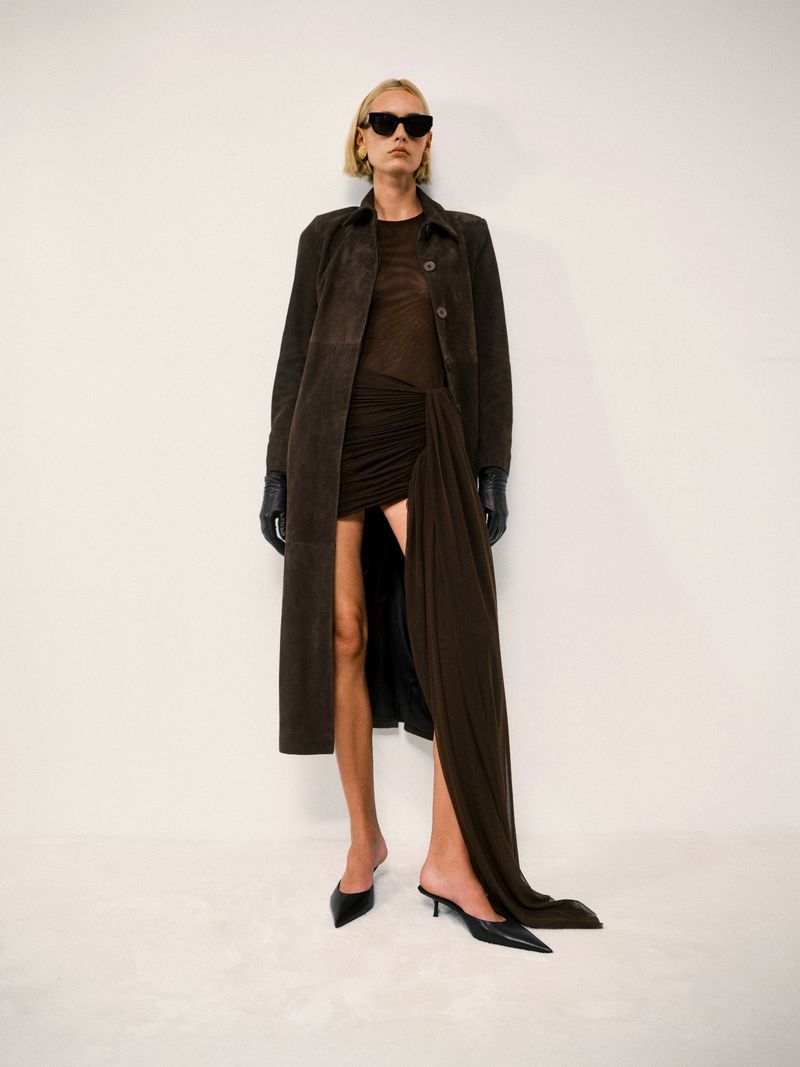 A woman poses in a brown coat, dark dress, sunglasses, and heels against a white backdrop, showcasing a minimalist, fashion-forward aesthetic.