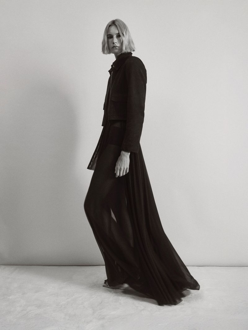 Monochrome fashion shot of a woman in a cropped jacket and sheer pants, posing in a photo studio with neutral background and textured floor.
