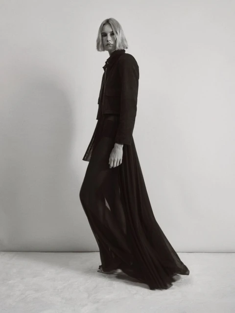 Monochrome fashion shot of a woman in a cropped jacket and sheer pants, posing in a photo studio with neutral background and textured floor.