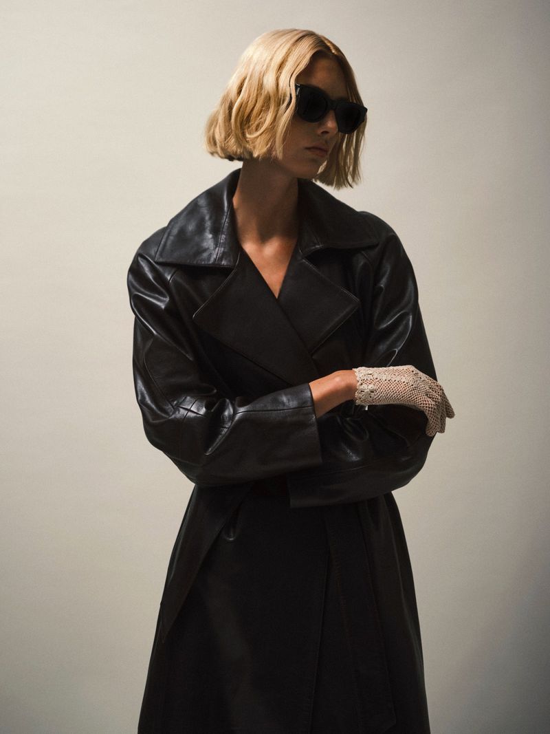 Blonde model in a black leather trench coat and vintage gloves poses against a neutral backdrop wearing dark sunglasses in a studio.
